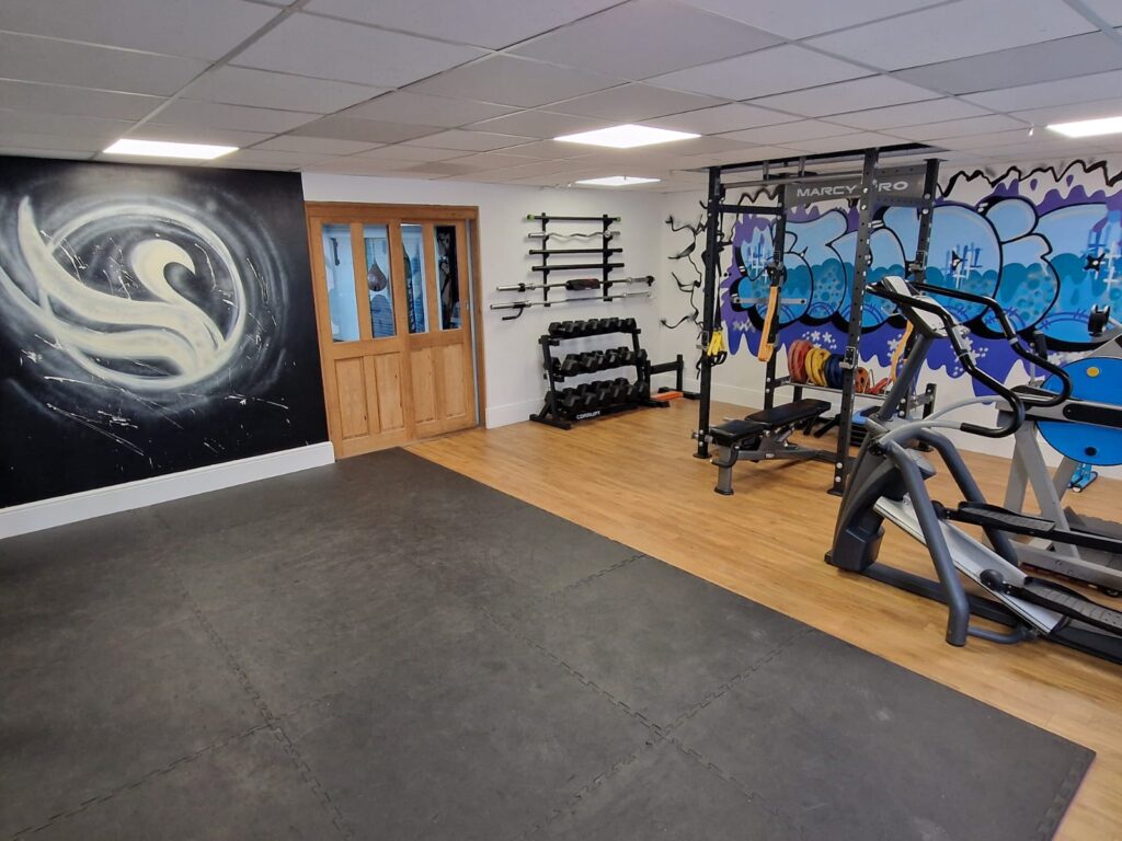 An image of the local gym, with a black mats, and the exercise equipment. Swansea Personal Training Logo is on the wall.