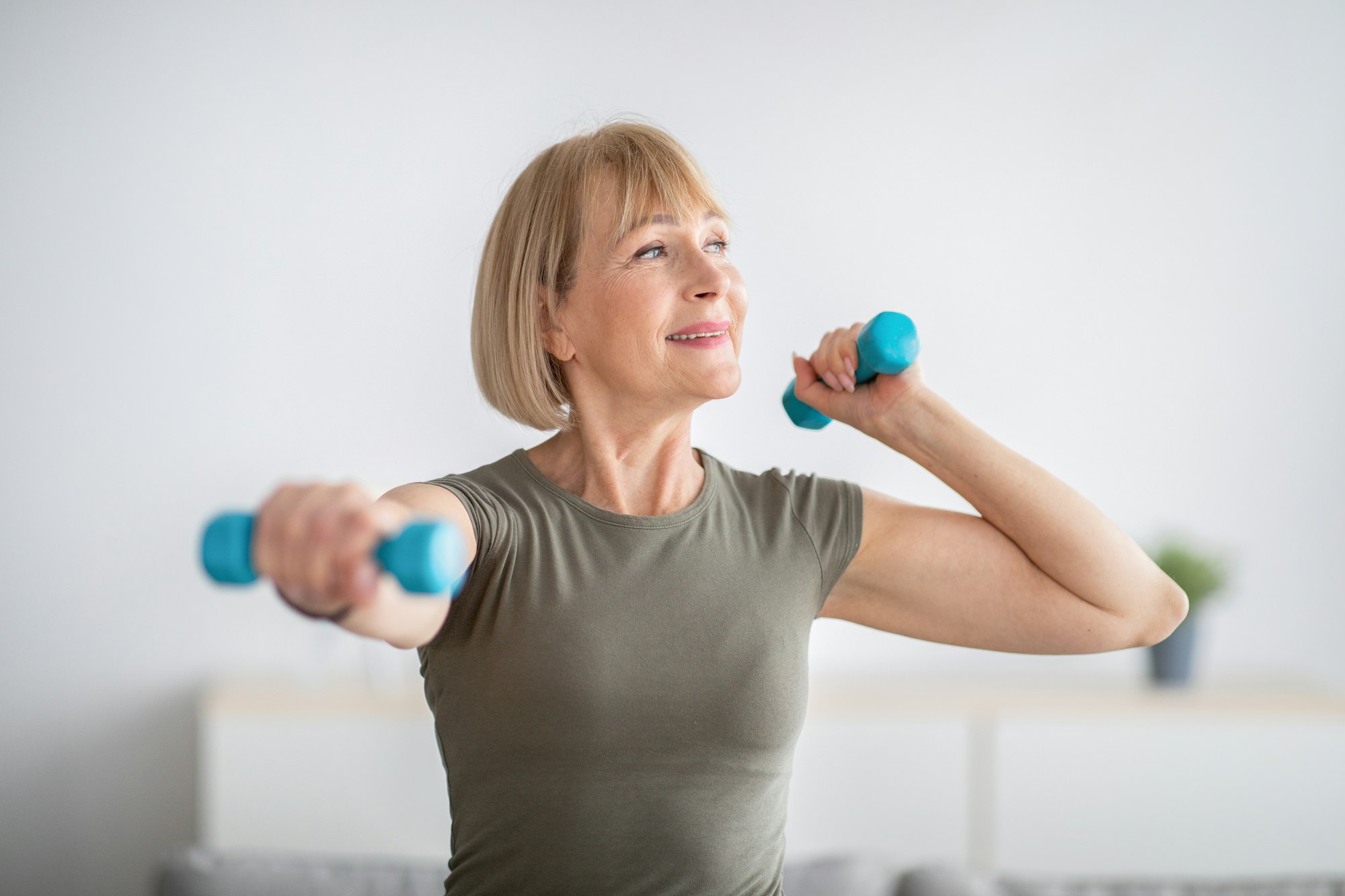 Domestic sports training. Positive senior woman doing exercises with dumbbells, strengthening her